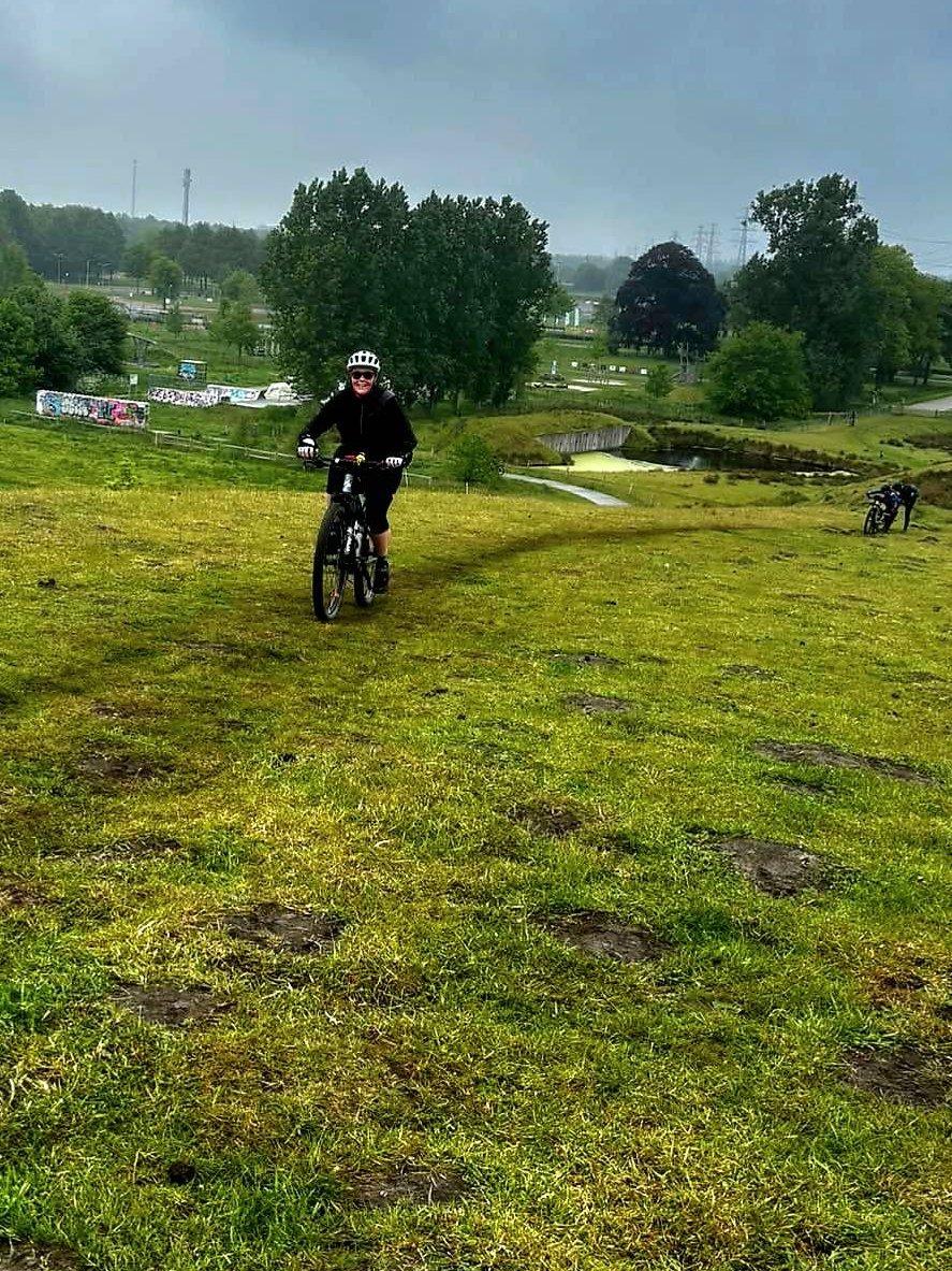 Lachende mountainbikester fietst over een grastalud onder een grijze lucht bij Assen